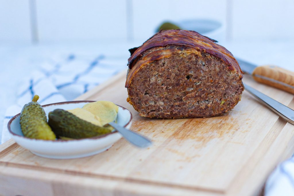 Omas Hackbraten mit Bacon obendrauf, aufgeschnitten auf einem Brett. Daneben Essiggurken und Senf.