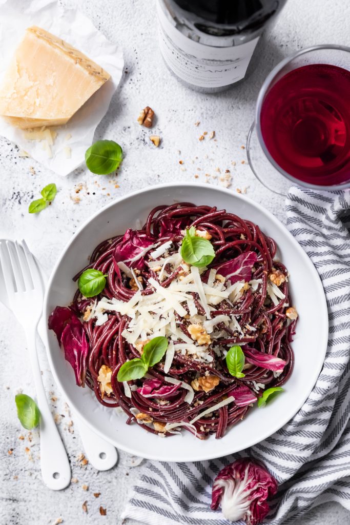 rote Spaghetti in einem weißen Teller, daneben ein Stück Parmesan und ein Glas Rotwein, von oben fotografiert