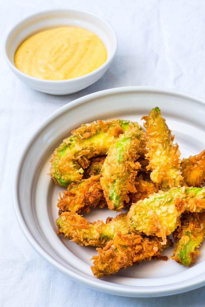 Frittierte Avocado Ecken auf einem weißen Teller, daneben ein gelber Dipp