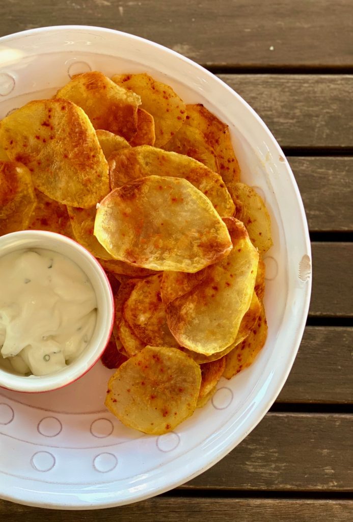 Selbstgemachte fettarme Kartoffelchips auf einem hellen Teller, mit einem Dip im roten Schälchen daneben, von oben fotografiert 