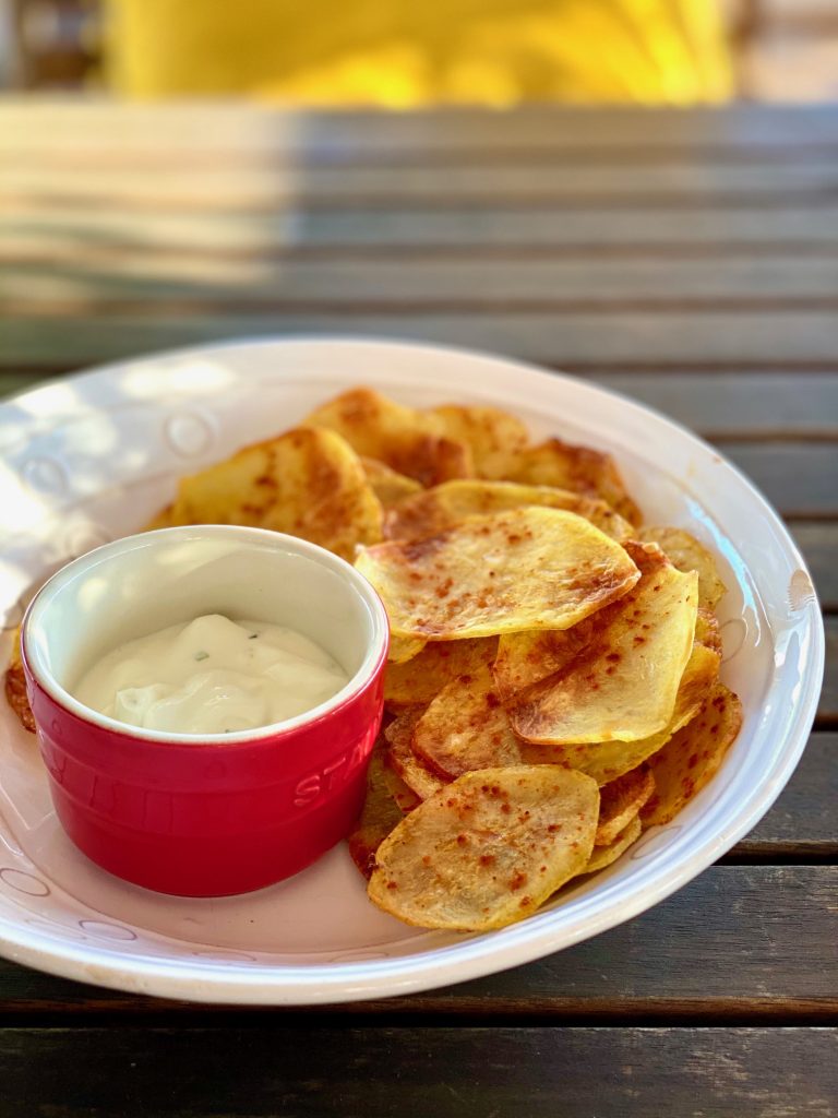 Selbstgemachte fettarme Kartoffelchips auf einem hellen Teller, mit einem Dip im roten Schälchen daneben 