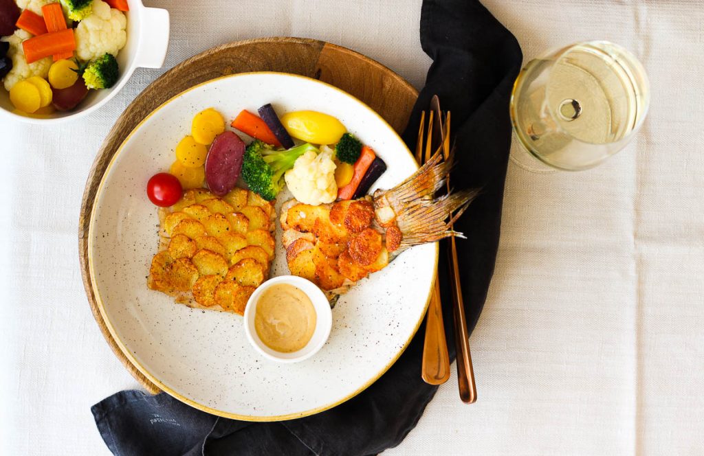 Karpfenfilet mit Kartoffelschuppen von oben fotografiert, daneben ein Glas Silvaner 