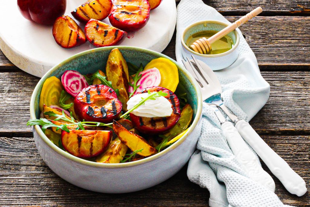 Bunter Salat mit gegrillten Pflaumen und Frischkäse dahinter ein weißes Brett mit Pflaumen