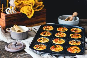 Beste Mini-Käse-Quiches mit Mangold in einem Blech, daneben ein Salztiegel aus Keramik und eine Schale mit Körnern